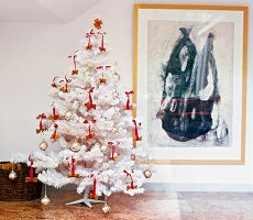 White Christmas tree decorated with baubles and red bows in front of wall painting