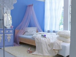 Bed with mosquito net, white blanket and cushions against blue wall in bedroom