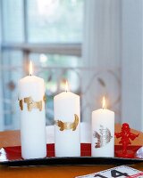 Drei weiße Kerzen mit goldenem Motiv auf einem roten Tablett.