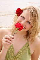 Portrait of beautiful woman wearing green top standing on beach and smelling red carnation