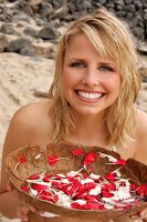 Pretty blonde woman sitting and holding bowl of white and red petals on beach, smiling