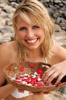 Pretty blonde woman sitting and holding bowl of white and red petals on beach, smiling