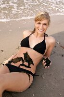 Portrait of pretty woman in black bikini lying on beach with seaweed on her belly, smiling