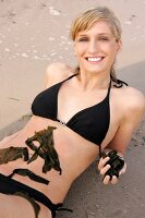 Portrait of pretty woman in black bikini lying on beach with seaweed on her belly, smiling