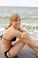 Portrait of blonde woman in bikini sitting and rubbing arm with sponge at beach, smiling