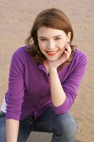 Portrait of beautiful woman wearing purple sweater and jeans sitting on beach, smiling