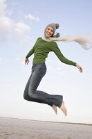 Beautiful woman wearing green sweater, jeans and cap jumping in air, smiling widely