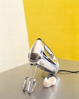 Plastic chromed hand mixer and eggshell on white-yellow background