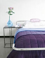 Close-up of bed with purple blanket and side table with blue flower vase