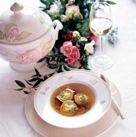 Suppe auf einem Teller, daneben eine Suppenterrine, Weinglas u. Blumen.