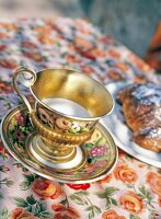 Close-up of golden cup and saucer with rose prints on floral patterned tablecloth