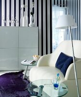 White armchair, glass table, lamp and wardrobe in front of striped wall