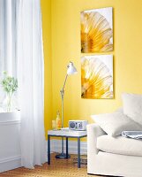 Weißes Sofa vor einer gelben Wand an der Blumenbilder hängen