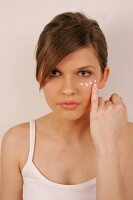 Portrait of pretty woman with brown hair applying cream below her eye with finger