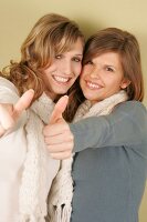 Two beautiful woman in sweater and scarf standing side by side showing thumbs up, smiling