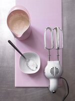 Bowl of cream with sugar and blender on pink board to prepare meringue