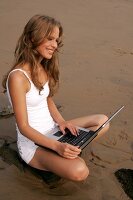 Pretty blonde woman in white outfit sitting on stone with laptop on lap at beach, smiling