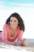 Portrait of pretty woman with windswept hair wearing pink sweater lying on sand, smiling