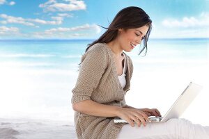 Frau sitzt am Strand und schreibt auf ihrem Laptop