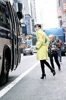 Woman wearing long coat and high heels walking on street along bus, looking over shoulder