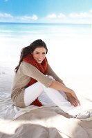 Portrait of woman in beige cardigan and red scarf, sitting on beach and smiling