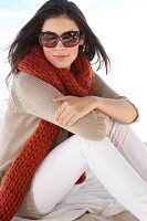 Beautiful woman wearing sunglasses, red scarf and beige cardigan sitting on beach, smiling