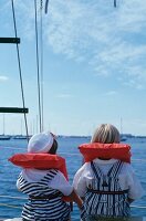 2 Kinder mit Schwimmwesten sitzen auf einem Segelboot, Rückansicht.