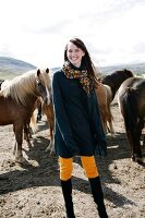Happy woman wearing poncho and scarf standing in front of icelandic horses, laughing