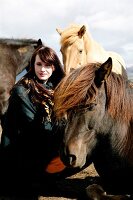 Beautiful brown haired woman wearing poncho and scarf crouching next to iceland horses