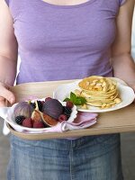 A woman holding a tray of pancakes and a fruit platter
