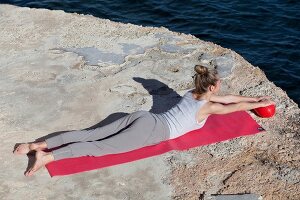 Woman lying on front and holding ball while performing workout for lower back near sea