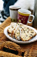 A plate of stollen cake and cups of hot drinks on top of a picnic basket in the open air