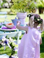 Mädchen im rosa Kleid & mit Blumenkranz am Tisch im Garten