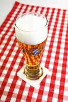 A glass of beer with a head of foam on a red and white checked tablecloth