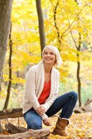 Pretty blonde woman wearing white sweater crouching and smiling in autumn forest