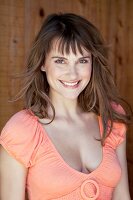 Portrait of beautiful brown haired woman in orange top in front of wooden wall, smiling
