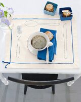 A cup of coffee with biscuits on a decorative place mat with outlines of cutlery and decorative ribbon