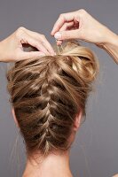 Rear view of blonde woman fixing hairpin in plaited hair to prepare updo, close-up