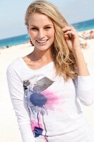 blonde Frau in weißem Shirt am Strand, blickt lächelnd in Kamera