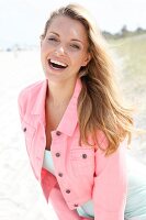 blonde Frau am Strand in rosa Kurzjacke, lacht, hat Spaß