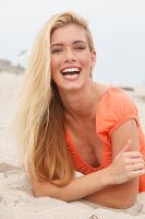 Portrait of happy blonde woman with long hair wearing orange top lying on beach, smiling