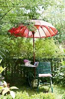Sitzplatz in sommerlichen Garten mit Gartenstuhl, Gartentisch & rotem thailändischen Sonnenschirm