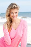 Portrait of pretty blonde woman wearing pink sweater sitting on beach, smiling