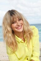 blonde Frau in gelber Bluse am Strand, lacht in die Kamera