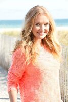 Blonde woman in an orange sweater on the beach, smiling at the camera
