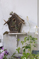 Vogelhaus aus Naturmaterialien auf Regalbrett, umgeben von Anemonen und Petunien im Garten