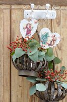 Körbe mit Hagebuttenzweigen und Blättern sowie Herzen mit Weihnachtsmotiven hängen auf Haken an einer Holzwand