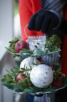 Weihnachtsgesteck auf einer Etagere mit Äpfeln, Tannenzweigen, Misteln, Moos, Windlicht und Christbaumkugeln