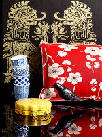Kissen mit floralem Bezug, Vogelfigur, Schatulle, Pappbecher vor asiatischem Wandbehang