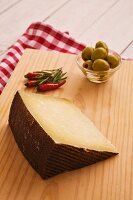 A wedge of Manchego, chillies and a small bowl of olives on a wooden board